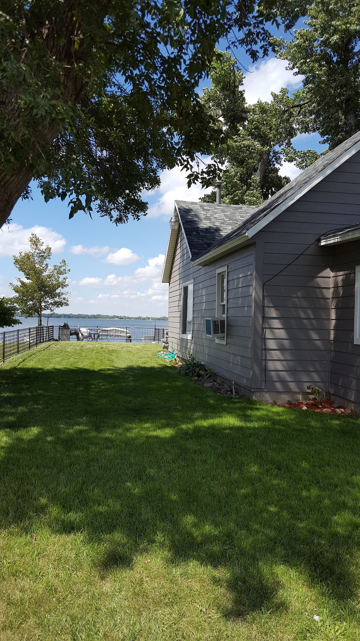 The exterior view of the lakefront cabin showcases a well-maintained grassy area adjacent to the structure, framed by trees. A glimpse of the lake can be seen in the background, with seating available along the shore, inviting relaxation and outdoor enjoyment.
