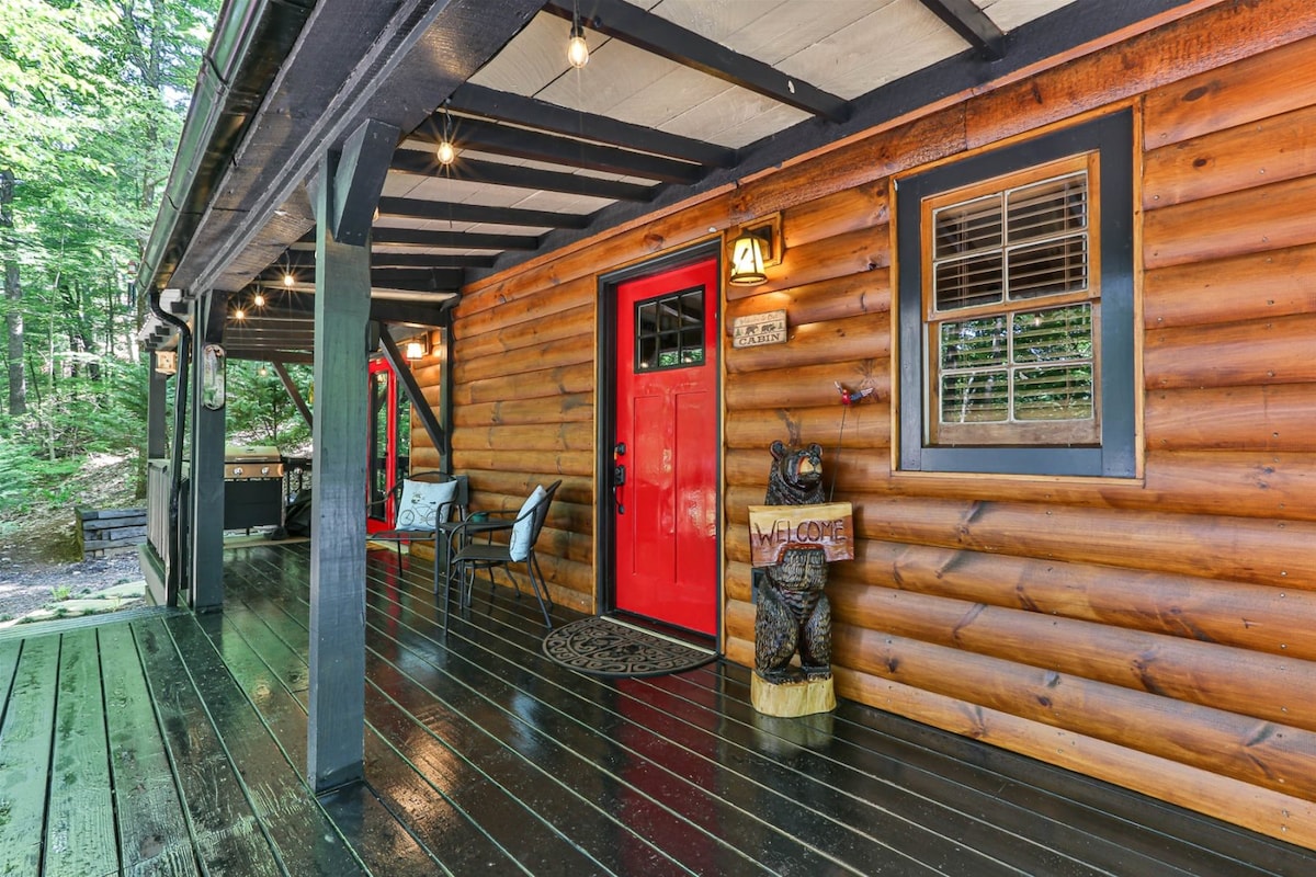 The entrance features a rustic wooden porch with dark flooring and logs supporting the roof. A bold red door welcomes guests, complemented by a charming bear statue on a pedestal. Two chairs sit nearby, providing a cozy spot to enjoy the natural surroundings.