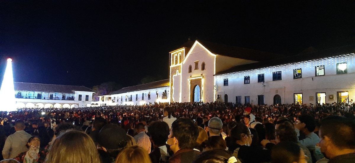 A lively crowd gathered in a spacious plaza during nighttime, illuminated by decorative lights. A grand colonial building stands in the background, featuring white walls and arched doorways. The atmosphere showcases a vibrant community gathering, with the architecture illuminated against the night sky.