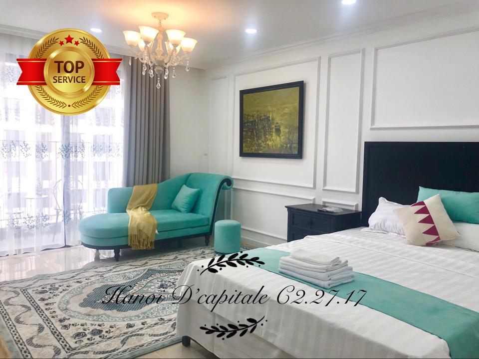 A well-appointed bedroom features a spacious bed with white linens and decorative pillows. A turquoise chaise lounge complements the room's color scheme and a chandelier adds elegance. Natural light filters through large windows with sheer curtains, illuminating the decorative area rug beneath.
