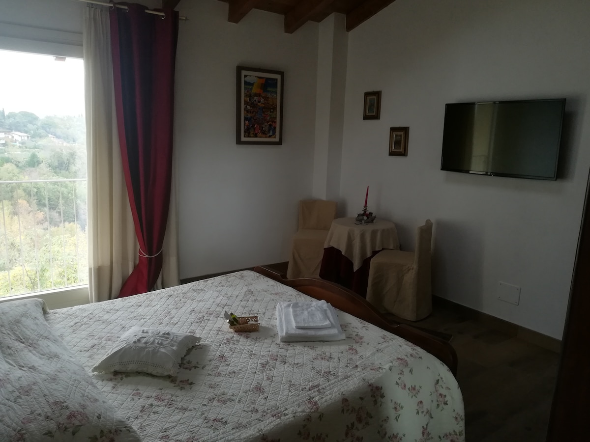 A spacious bedroom is presented, featuring a large bed with floral bedding and neatly arranged linens. A small dining table with two chairs occupies one corner, while a flat-screen television is mounted on the wall. Natural light enters through a window with red drapes, offering a serene view.
