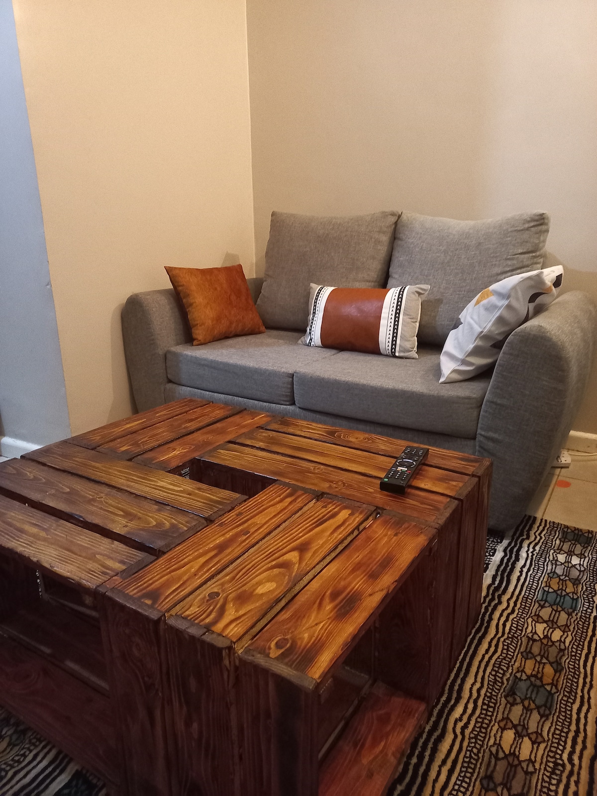 A cozy seating area features a simple gray sofa adorned with two decorative pillows. A rustic wooden coffee table, constructed from crates, adds an element of charm. A woven rug lays beneath, enhancing the inviting environment.