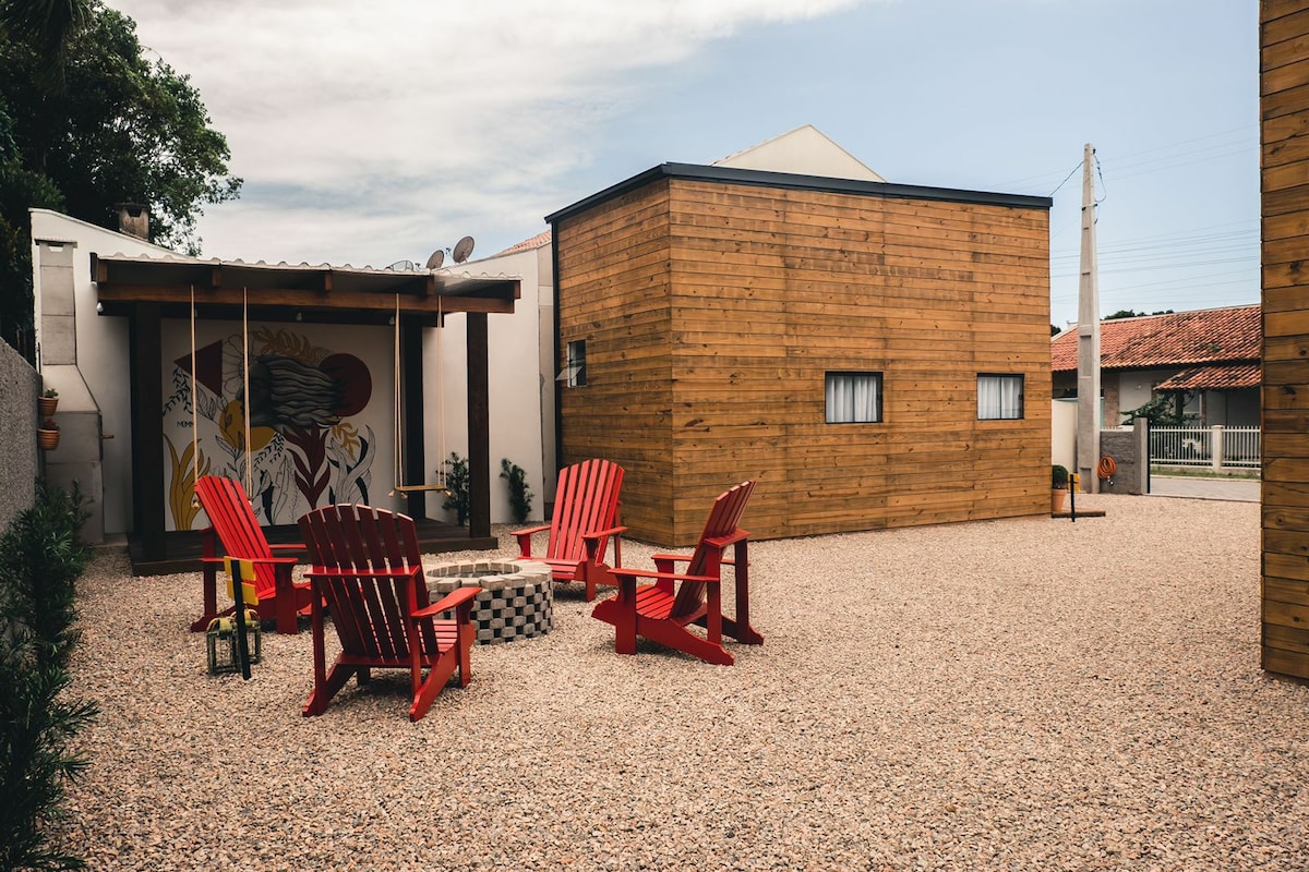 An outdoor area features a circular arrangement of red Adirondack chairs surrounding a stone fire pit. A small wooden structure with windows and a welcoming mural is visible in the background, complemented by a gravel surface that enhances the spaciousness of the setting.