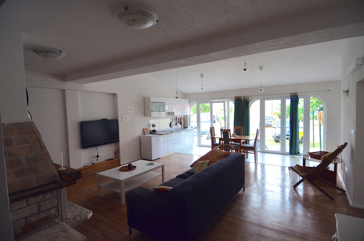 An open-plan living and dining area is shown, featuring a spacious sofa and a coffee table. Large glass doors provide natural light and access to the garden. A television is mounted on the wall, with a fireplace adding character to the space. Wooden floors enhance the inviting atmosphere.