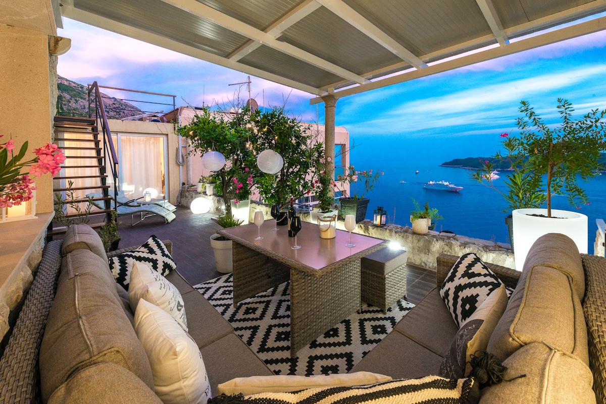 A terrace is furnished with a lounge sofa and a large table, surrounded by decorative plants and soft lighting. An expansive view of the Adriatic Sea is visible, with boats dotting the water and a staircase leading to another level.