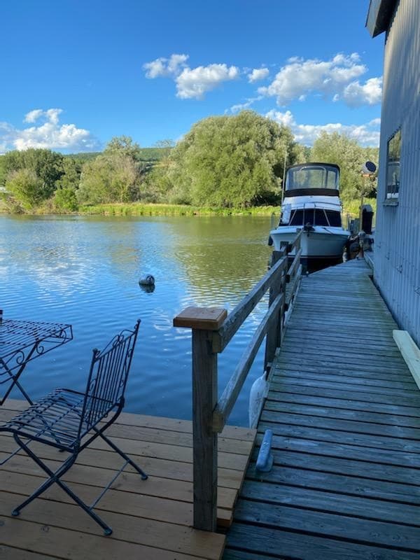A wooden walkway leads to the water's edge, featuring a small dining table with two chairs. A boat is moored nearby, and reflections of trees and clouds can be seen on the calm surface of the canal, offering a serene view.