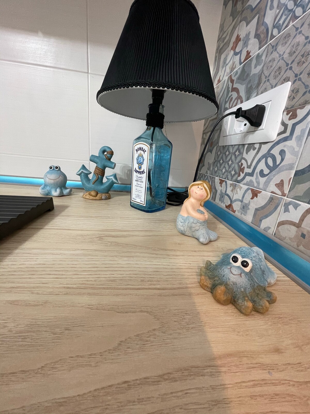 A kitchen countertop is featured, displaying a lamp with a pleated black shade next to decorative figurines. A blue decorative bottle is positioned alongside a small anchor figure and a whimsical creature, creating a playful yet functional space.