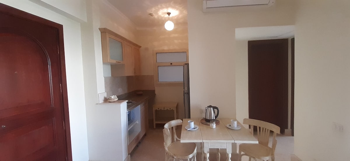 A well-equipped kitchen area is visible, featuring wooden cabinets and stainless steel appliances. A small dining table with two chairs is positioned against the wall, complemented by a kettle and two cups. Soft lighting is provided by a ceiling fixture.