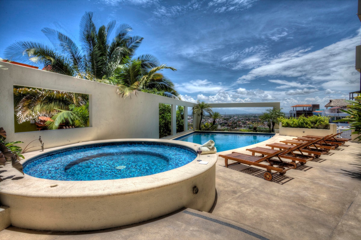 An outdoor area features a large swimming pool alongside a circular hot tub. Sun loungers are arranged around the space, with lush palm trees providing a green backdrop. Expansive views of the surrounding area and a clear sky are visible, enhancing the tranquil setting.