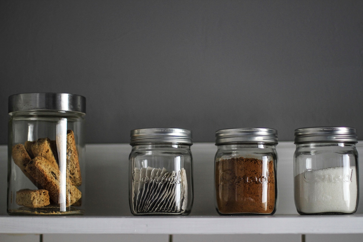 Glass jars are organized on a shelf against a gray wall. One jar contains baked goods, while three smaller jars hold sugar, cocoa powder, and tea bags, respectively. The arrangement offers an organized and minimalist touch to the kitchenette.