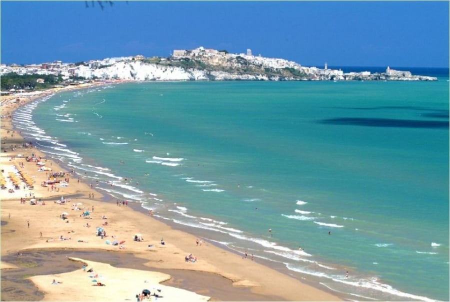 The image captures a sandy beach curving along a vibrant turquoise sea, dotted with sunbathers and umbrellas. In the background, a coastal town is visible, with white buildings and a fortress atop a hill under a clear blue sky.