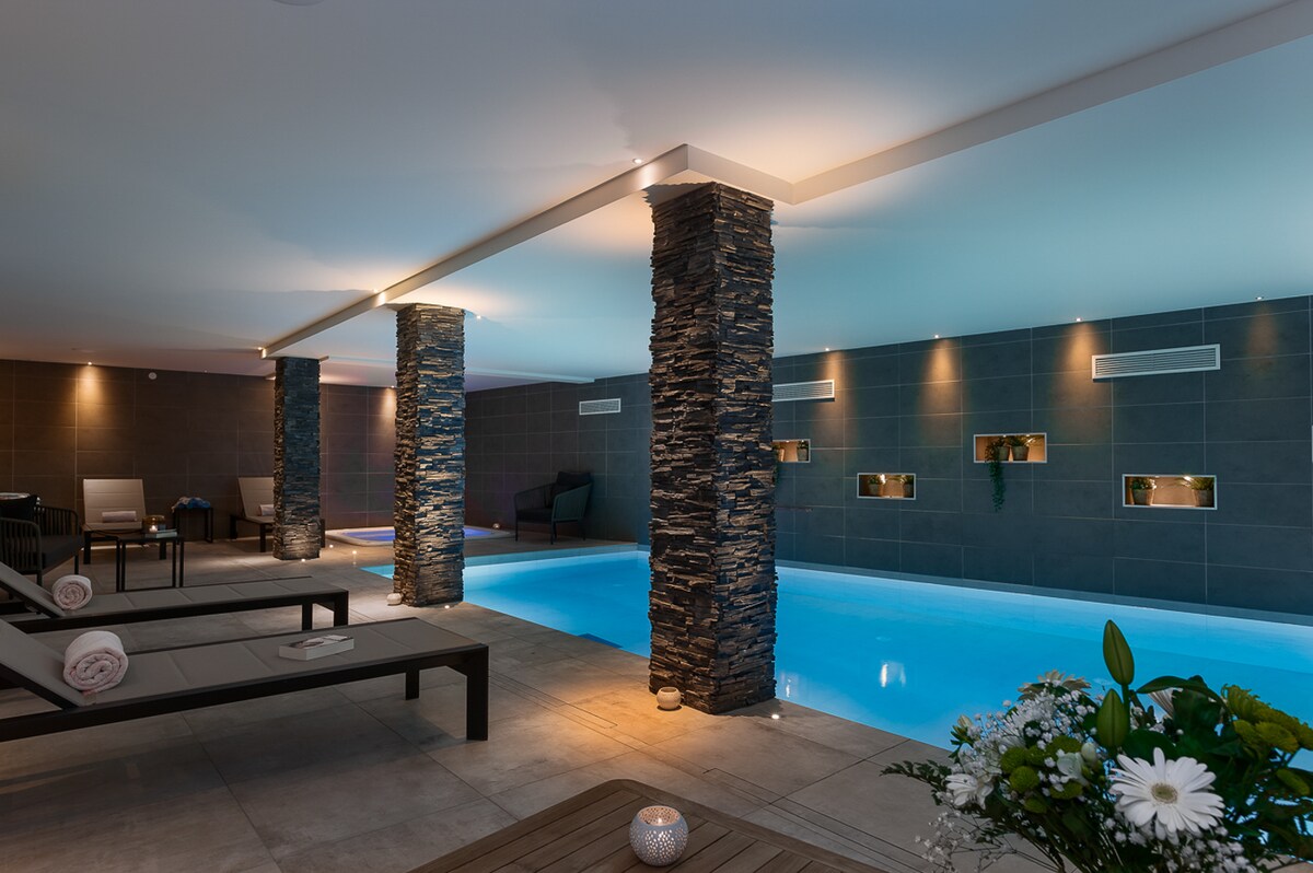 An indoor swimming pool area is illuminated with soft blue lighting, highlighting the clear water. Stone pillars are positioned along the sides, and comfortable lounge chairs with rolled towels are arranged nearby. A small table holds a decorative candle and a bouquet of flowers.