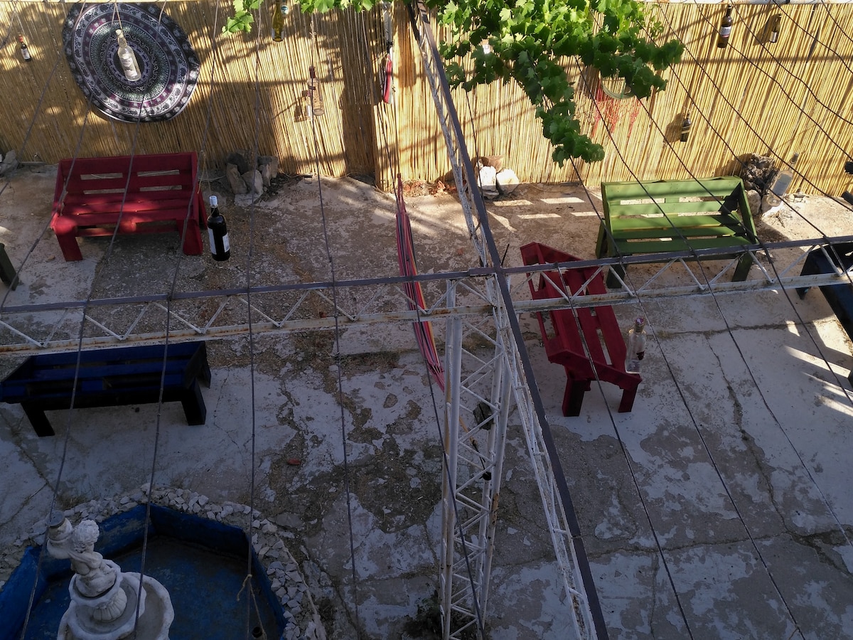 An aerial view of a serene outdoor space showcases a circular fountain surrounded by colorful seating areas. Wooden benches in red and green are arranged on the sandy ground, with a shaded area formed by overhead vines and decorative elements offering a relaxed setting.