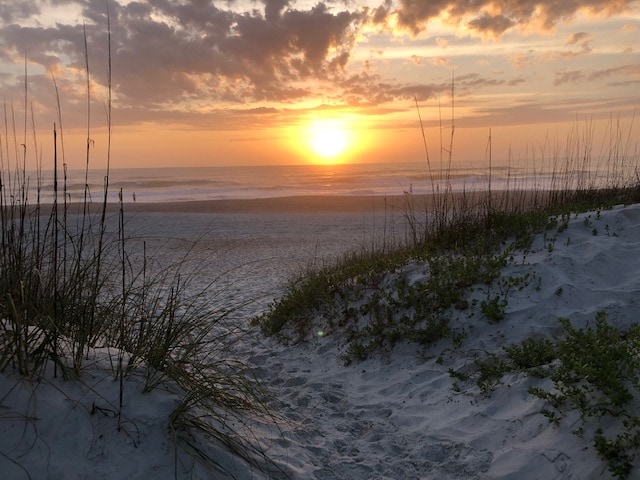 A serene beach view captures the sun setting over the horizon, illuminating the ocean with warm golden hues. A sandy path leads through grassy dunes, highlighting the natural beauty of the seashore. Soft clouds scatter across the sky, enhancing the peaceful atmosphere.