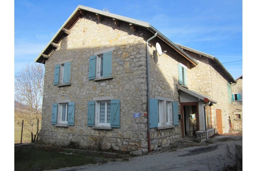 A charming two-story stone house is depicted with a stone exterior and blue shutters. Large windows allow natural light to fill the interior. The entrance is framed by a small porch, while a gravel path leads to the front door, surrounded by a scenic rural landscape.