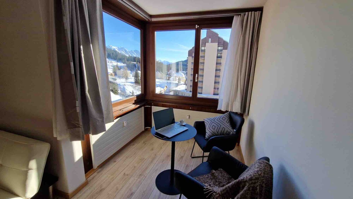 A cozy corner features large windows that offer a scenic view of snow-covered mountains. A small round table is paired with two armchairs, creating an inviting space for relaxation or work. Natural light fills the room through the expansive glass.