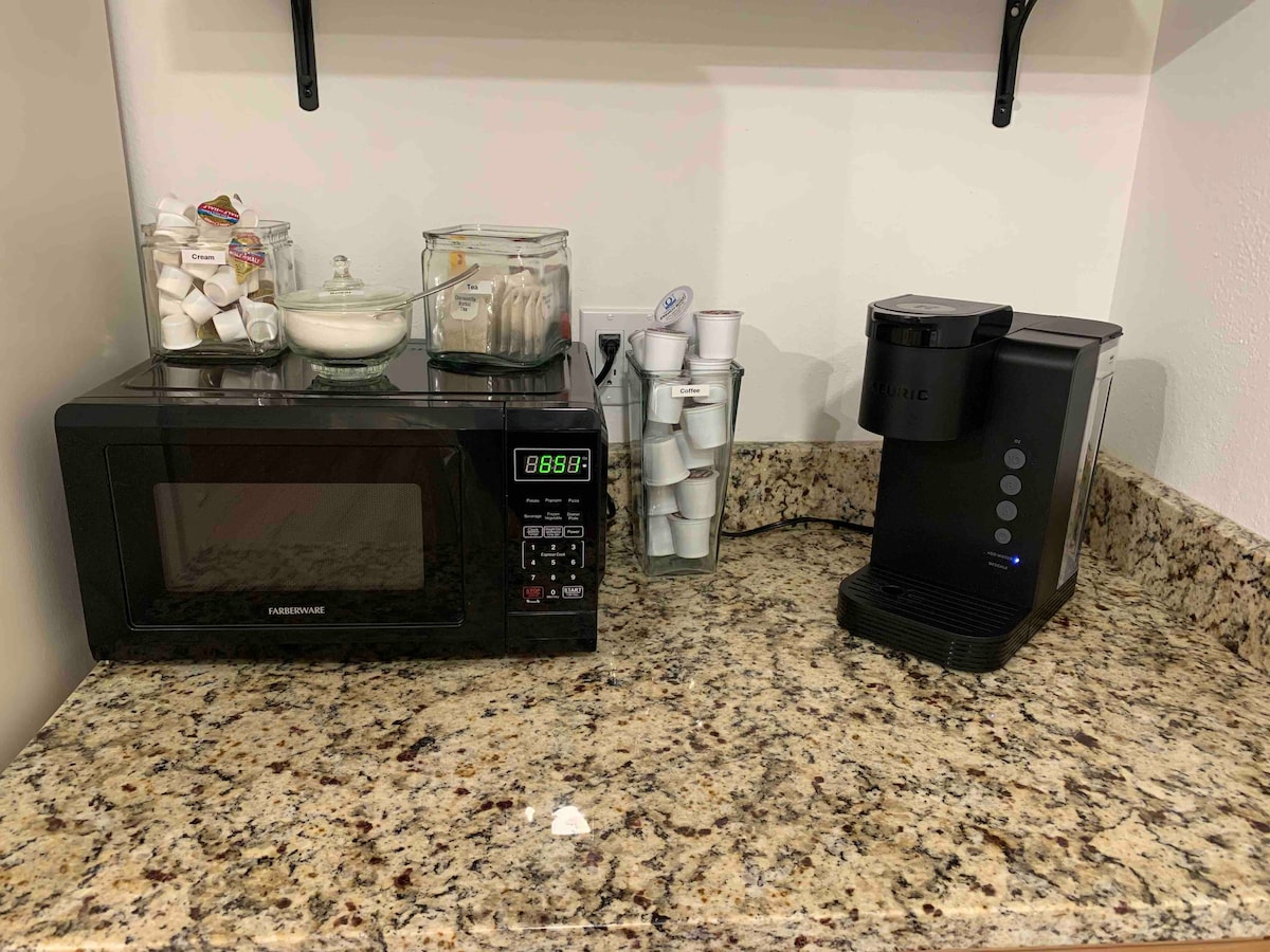 A kitchen countertop features a microwave and a coffee maker, both positioned beside a variety of coffee pods. A glass container holds sugar packets, while additional items are neatly organized in a dish. The granite surface adds a natural texture to the space.