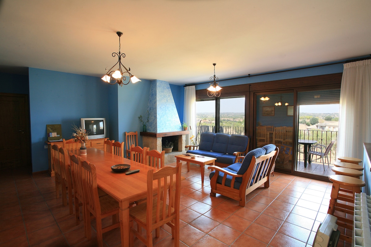 A dining area with a large wooden table is presented, surrounded by matching chairs, featuring a cozy fireplace built into a blue stone wall. A blue sofa and additional seating are visible in a well-lit space, with panoramic windows revealing a scenic view.