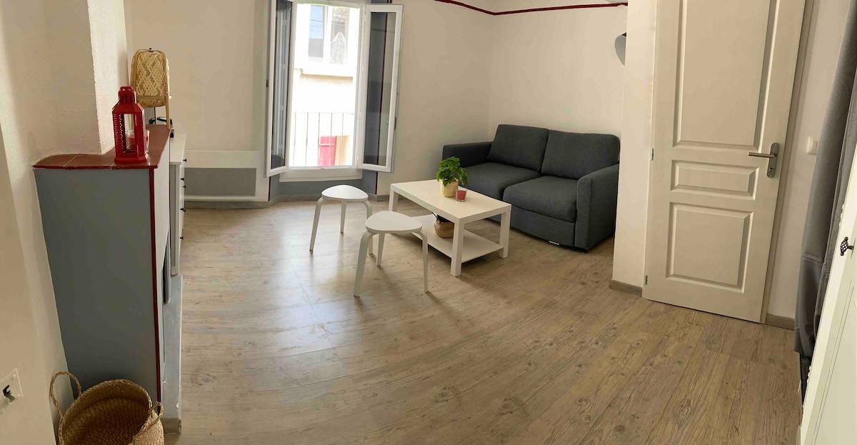 A bright living space features a gray sofa positioned next to a white coffee table, complemented by a small side table. Large windows allow natural light to enter, creating a airy atmosphere. Light-colored wooden flooring enhances the open feel of the area.
