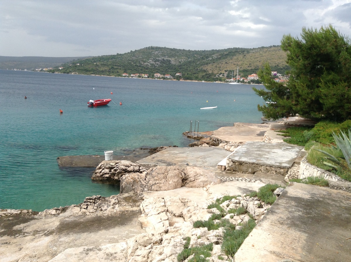 A serene coastal area showcases calm turquoise waters, with a rocky shoreline leading toward the horizon. A small red boat is anchored nearby, while gentle waves lap against the stone pier. Lush green hills rise in the background, accentuating the peaceful setting.
