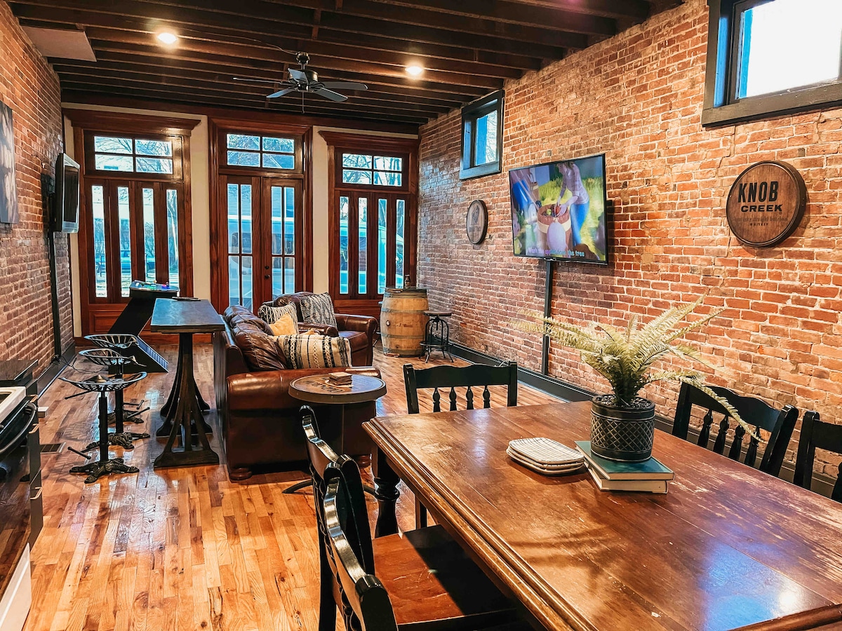 An inviting open living space showcases exposed brick walls and wooden beams. A comfortable leather sofa faces a large television, while a dining table with chairs occupies one corner. Natural light streams through large windows, enhancing the warm atmosphere of the room.
