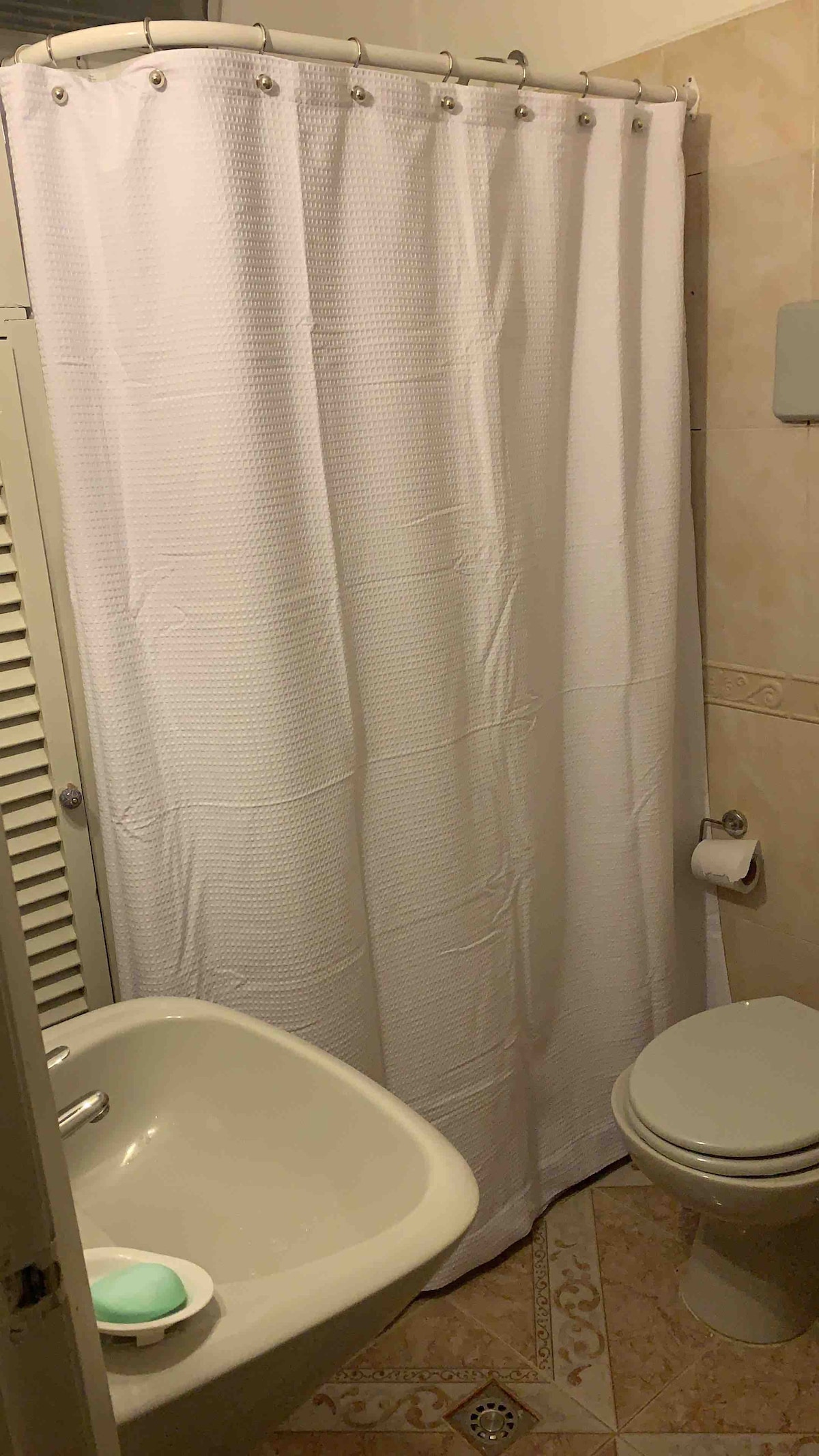 A compact bathroom is equipped with a pedestal sink and a toilet. A white shower curtain offers privacy while partially concealing a shower space. Neutral tiles are seen on the floor, enhancing the room's simplicity.