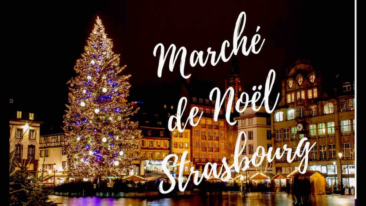 A festive Christmas market scene in Strasbourg is captured at night. A large, brightly lit Christmas tree adorned with blue and white ornaments stands prominently alongside historical buildings, which are softly illuminated, reflecting in the water below.