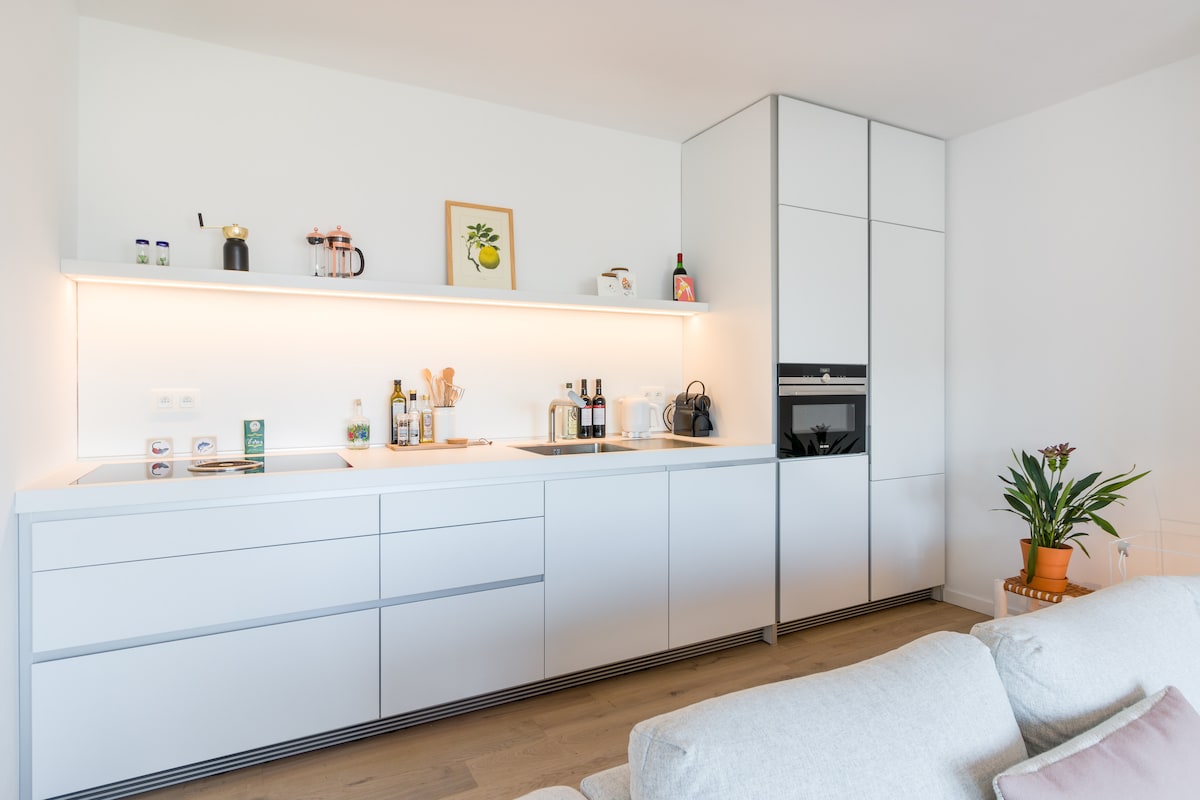 A modern kitchen features sleek white cabinetry and a countertop equipped with essential kitchen tools and cooking ingredients. A small artwork is displayed on the wall, and a potted plant adds a touch of greenery. Ample storage is provided by the built-in units.