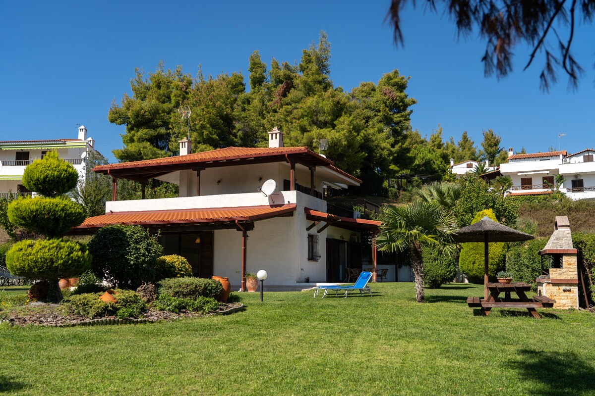 A spacious two-story house is set amidst a lush green garden, featuring manicured hedges and ornamental trees. An outdoor seating area with a table and benches is visible, along with a cozy barbecue area. Clear blue skies and sunlight create a bright atmosphere for relaxation.