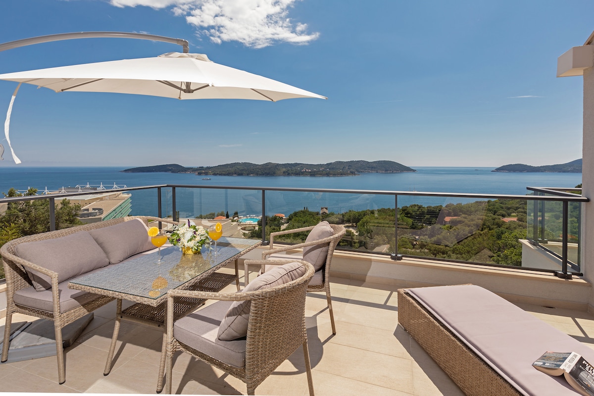 A spacious terrace features modern wicker furniture, including a table set with fresh fruit and drinks. An umbrella provides shade from the sun, while panoramic views of the sea and nearby islands are visible in the background.
