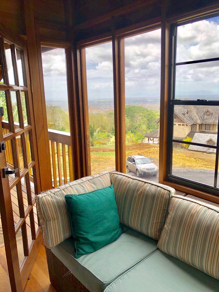A cozy seating area is framed by large glass windows, offering expansive views of the lush landscape and distant hills. A green cushion contrasts with the striped fabric of the sofa, inviting relaxation in the natural light that filters through the windows.