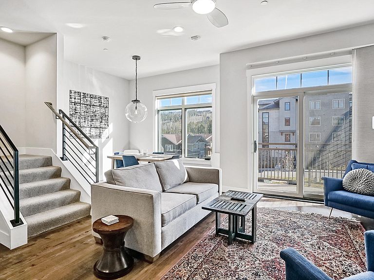 A cozy living area features a light sofa accompanied by a blue chair and a circular side table. Natural light fills the space through a large window with a view of the outdoor area. A staircase leads to the upper level.
