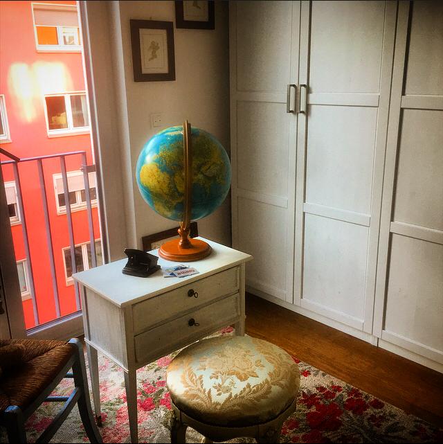 A small desk with a globe and a vintage telephone is positioned next to a floral-patterned stool. A white wardrobe offers storage, while large windows allow natural light to brighten the room, revealing a vibrant red building across the street.