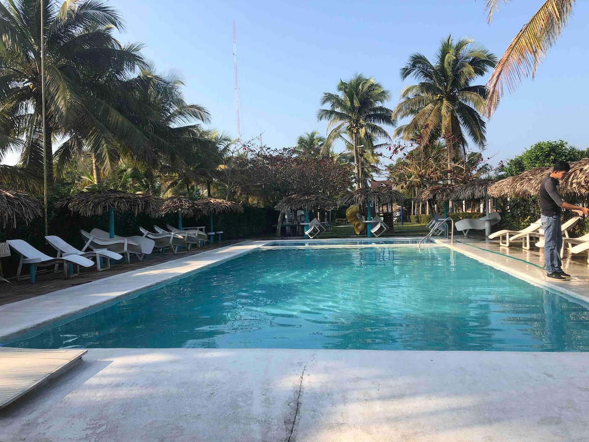 A rectangular swimming pool is surrounded by lush palm trees and thatched-roof cabanas. Sun loungers are positioned along the pool deck, inviting relaxation. The clear blue water reflects the sunlight, creating a calming atmosphere in a tropical setting.