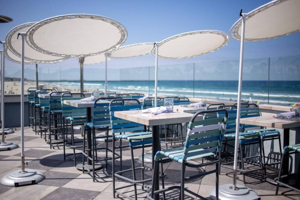An outdoor dining area is presented with multiple tables and chairs, featuring blue and silver color schemes. Large umbrellas provide shade, while the ocean waves and sandy beach are visible in the background, creating a relaxed seaside atmosphere.
