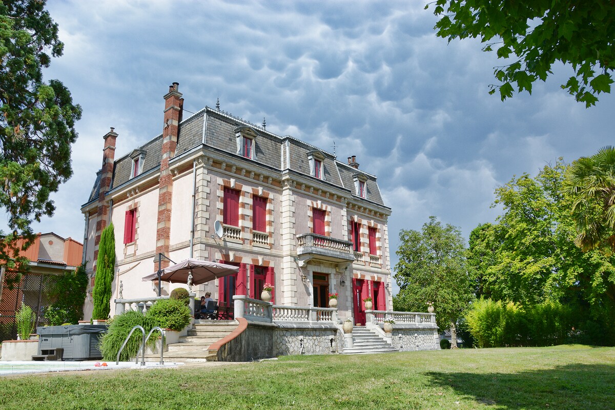 The chateau is depicted surrounded by lush greenery, featuring distinctive architectural details and large red-trimmed windows. A staircase leads to an inviting entryway, while a heated swimming pool is visible to the side. The scene is set under a dramatic sky, adding a captivating backdrop.