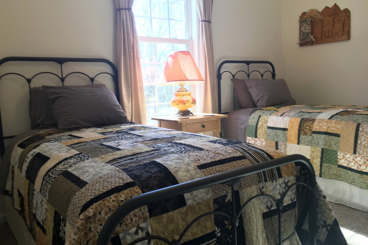 A bright and inviting bedroom is highlighted by two iron-framed twin beds, each covered with patchwork quilts in warm tones. A bedside table with a decorative lamp adds a soft glow, while natural light filters through the window dressed with light drapes.