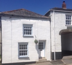 A charming cottage exterior is presented, featuring whitewashed walls and a textured slate roof. Two multi-paned windows are visible on the ground floor, complemented by a single door adorned with a small hanging plant. An archway is seen to the right, providing additional architectural interest.