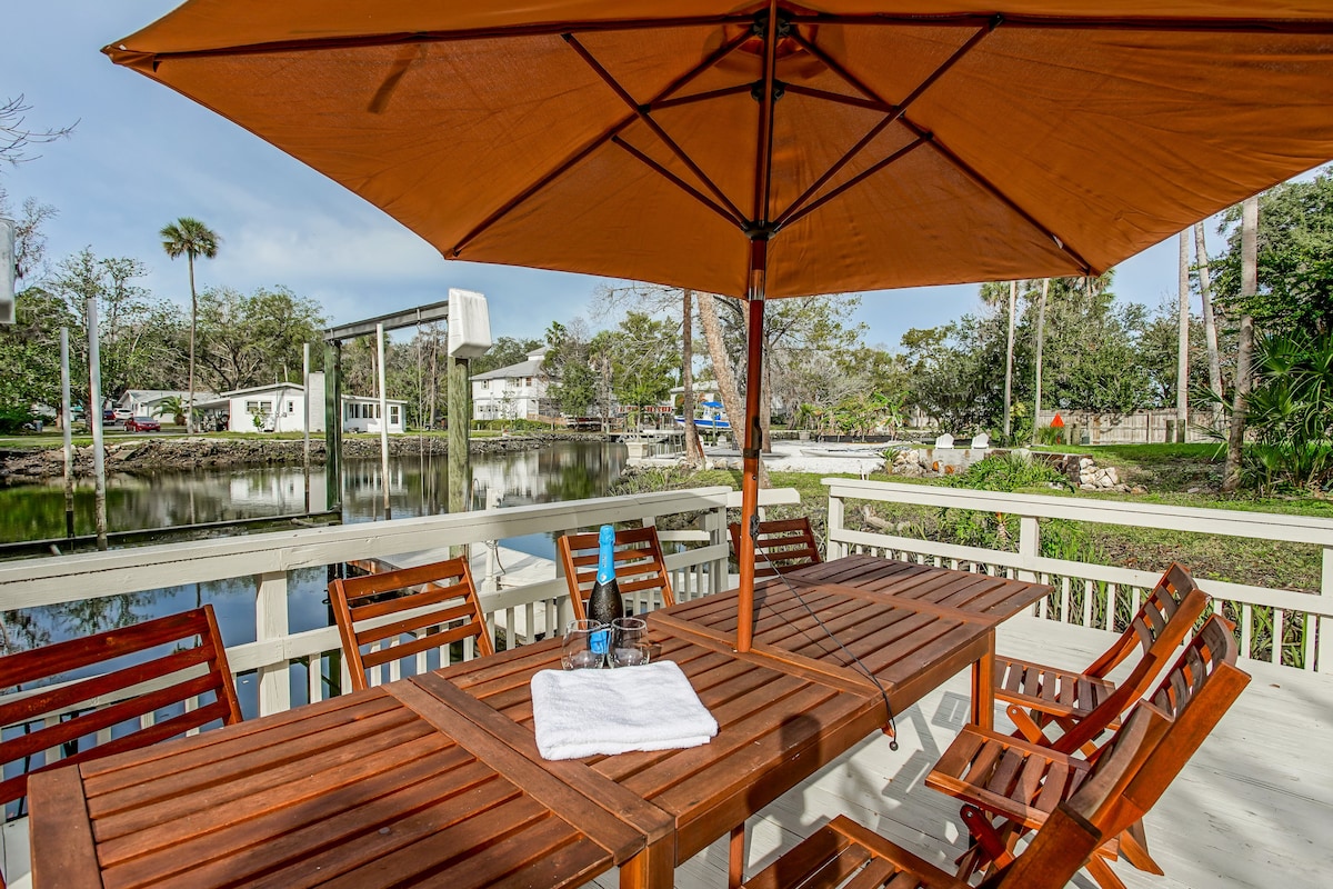 A spacious wooden deck is presented with an umbrella providing shade over a large table surrounded by multiple chairs. The tranquil canal reflects greenery and nearby structures, creating a serene outdoor space ideal for gatherings.