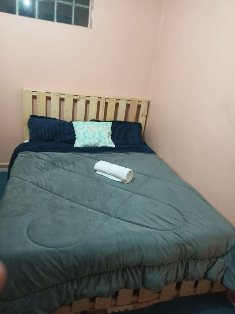 A compact sleeping area features a wooden bed frame with a plush gray blanket draped across the spacious bed. Two dark blue pillows are positioned at the headboard, while a rolled towel is placed centrally on the bed. A small window allows natural light to enter the room.