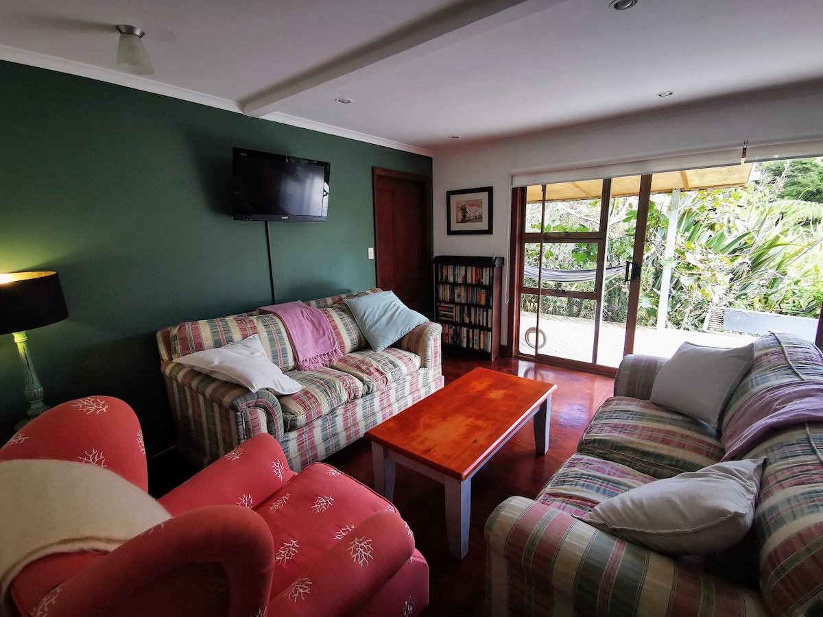 A cozy living area features a striped and solid pink sofa set, accompanied by a wooden coffee table. Natural light floods in from sliding glass doors, leading to an outdoor deck surrounded by lush greenery. A wall-mounted television adds to the convenient layout.