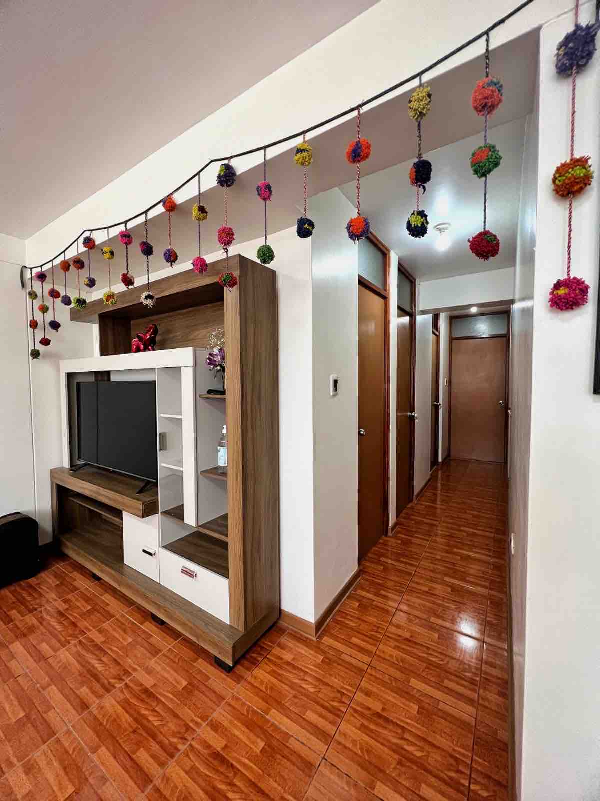 A modern entertainment unit is positioned against a wall, featuring a television and decorative elements. Colorful pom-poms are artistically displayed along the ceiling, adding a cheerful touch. A hallway leads to multiple doors, indicating access to private rooms or spaces.