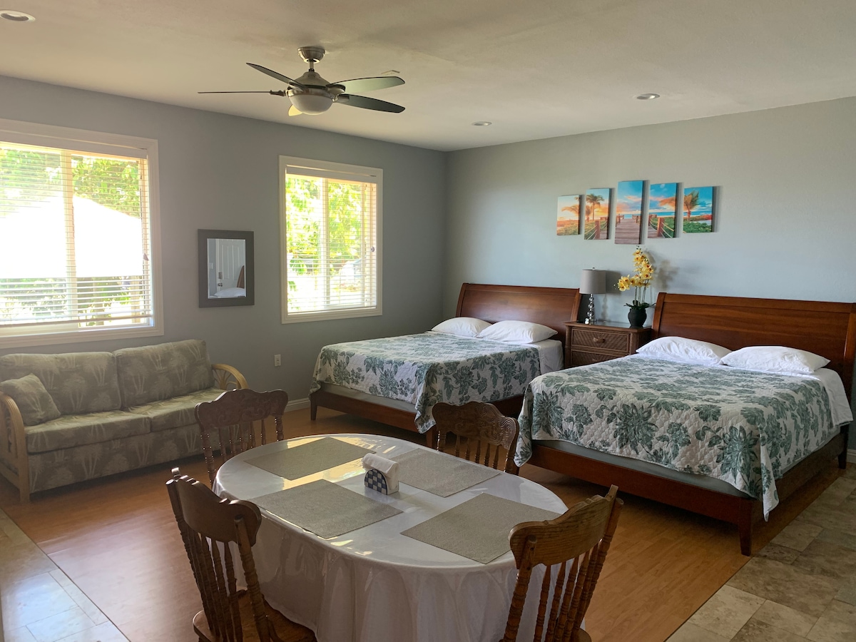 A spacious studio features two queen beds with floral-patterned bedding, a comfortable sofa, and a dining table with chairs. Natural light illuminates the room through multiple windows, offering views of the outdoor area.