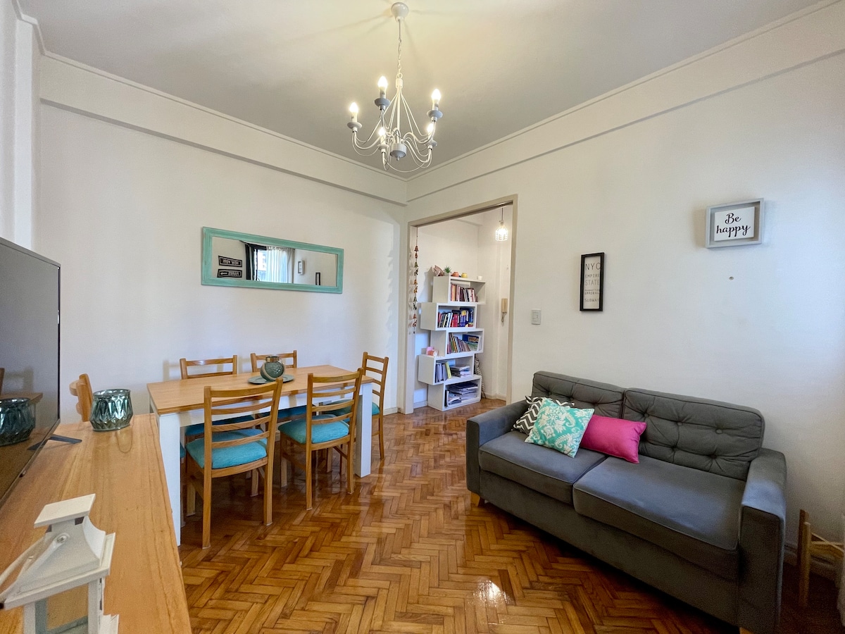 A comfortable living and dining area features a gray sofa adorned with a colorful pillow. A wooden dining table with six chairs is positioned nearby. Natural light streams through the windows, illuminating the polished wooden floor. A decorative chandelier hangs from the ceiling.