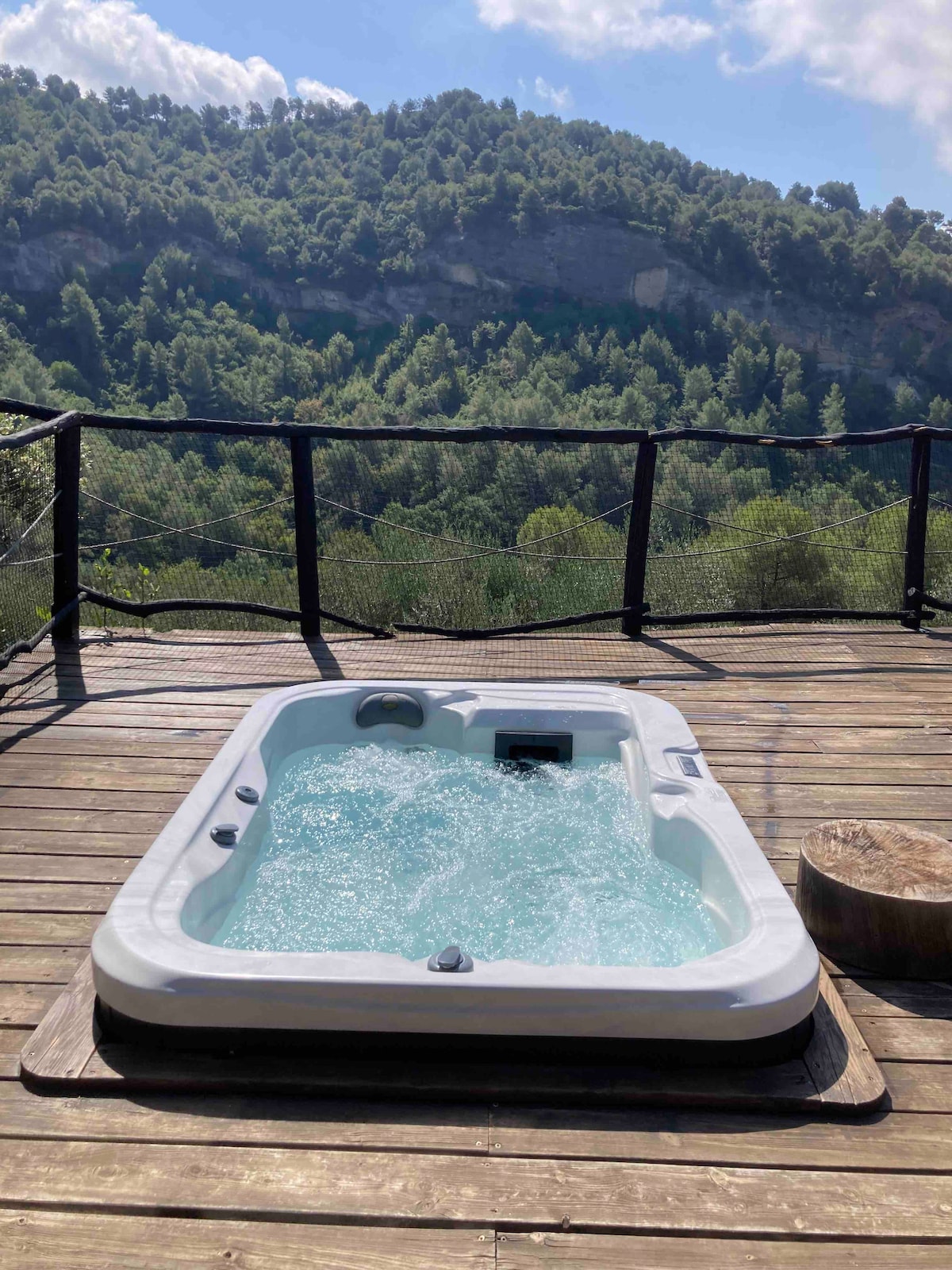 A private jacuzzi is set on a wooden terrace, surrounded by natural scenery. The water bubbles gently, and the backdrop features a lush hillside, enhancing the tranquil atmosphere of the outdoor space.
