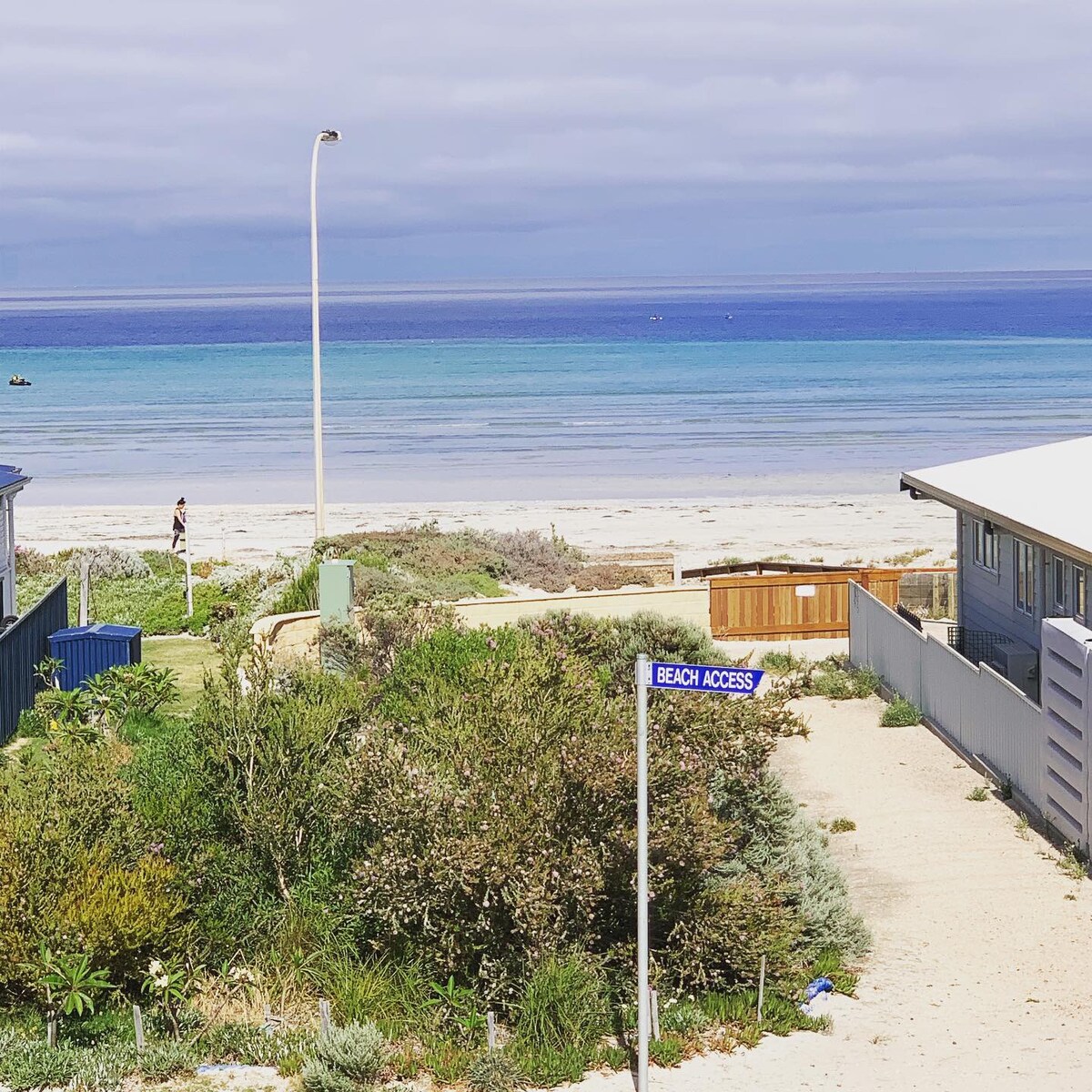 A clear view of the sandy beach is visible, with gentle waves lapping at the shore. A sign indicating 'Beach Access' stands prominently. Lush greenery lines the pathway leading toward the beach, complemented by a calm blue sea under a partially cloudy sky.