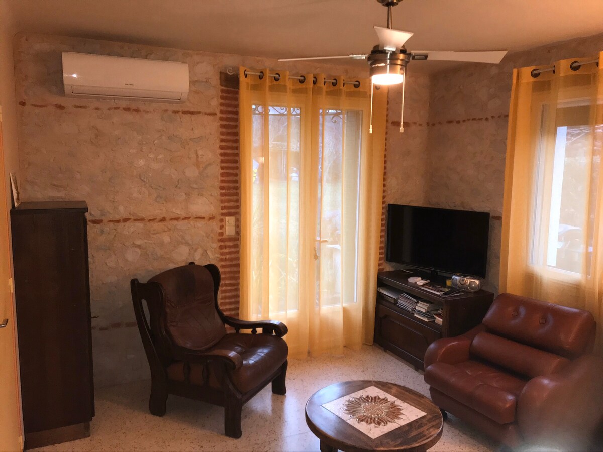 The living area features a blend of modern and rustic design with exposed stone walls and large windows draped in sheer yellow curtains. Comfortable seating includes a wooden armchair and a leather chair, with a small round table in the center. A TV is positioned against the wall.