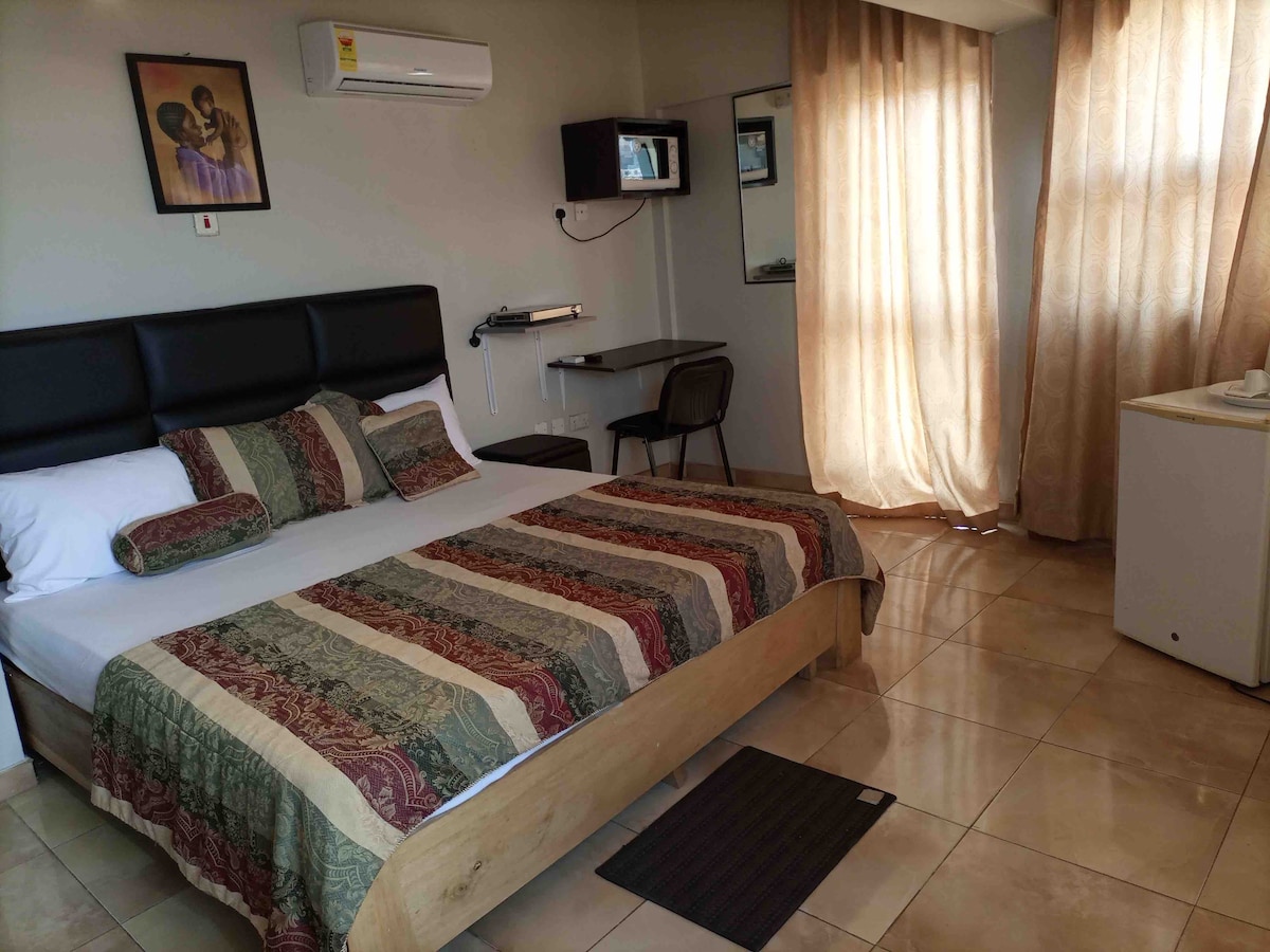 A spacious bedroom is furnished with a large bed adorned with a patterned blanket and decorative pillows. A work desk with a chair is positioned near a window draped with light curtains. A small fridge and wall-mounted television add convenience.