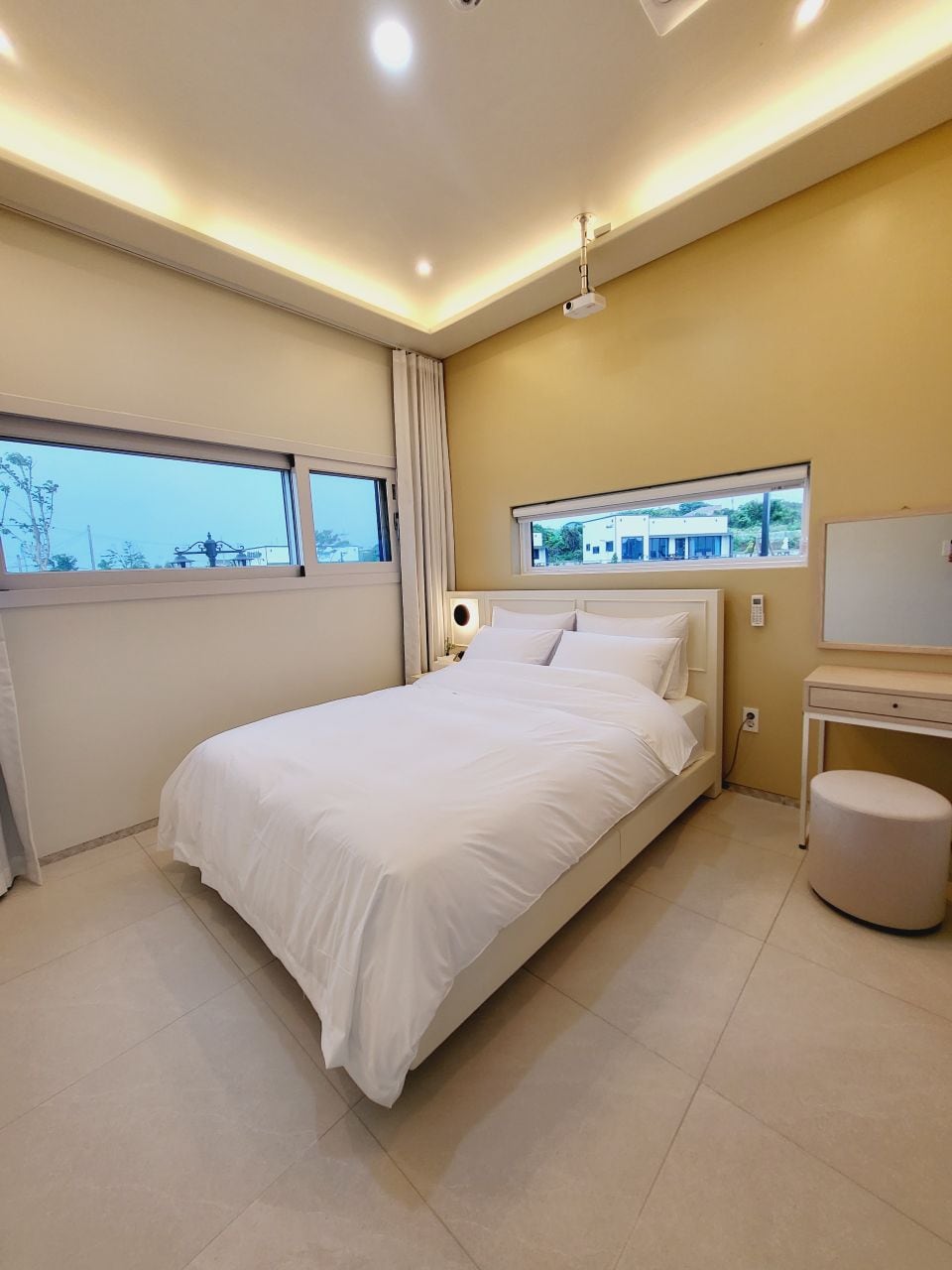 A simple bedroom features a spacious bed dressed in crisp white linens, complemented by soft lighting from recessed fixtures. Large windows allow natural light to fill the room, while a minimalist desk and a circular stool provide functional space without clutter.