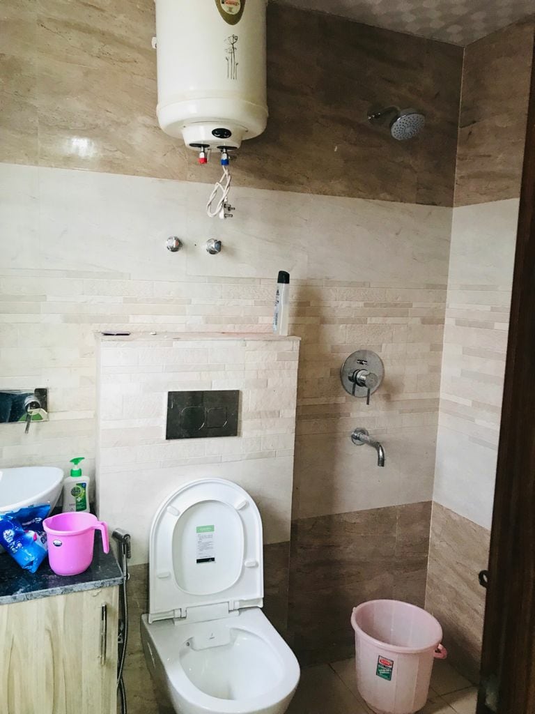 A clean and modern bathroom is equipped with a wall-mounted water heater and a showerhead. A toilet is positioned beside a sink with a small counter. Green and purple cleaning supplies are discretely arranged on the counter, and a pink waste bin is placed on the floor.